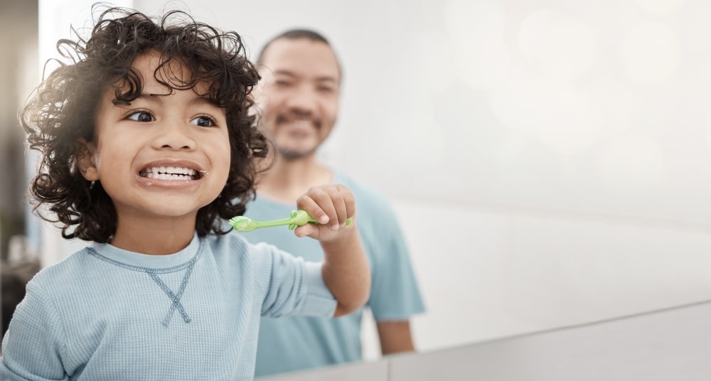 Jeune enfant qui se brosse les dents avec son père qui regarde à l'arrière